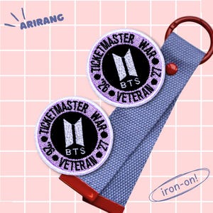 Op de afbeelding: Twee ronde patches met de tekst "TICKETMASTER WAR '26 VETERAN" en het BTS-logo. De patches zijn paars en zwart. Een lichtblauwe band met een rode accent en een metalen ring is aan de patches bevestigd. De tekst "iron-on!" is ook zichtbaar.