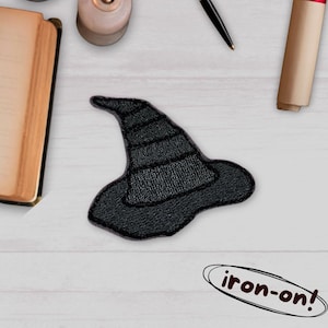 Könnte beinhalten: Schwarzer Bügelflicken in Form eines Hexenhuts. Der Hut ist mit horizontalen Linien versehen und hat eine geschwungene Krempe. Der Aufnäher befindet sich auf einer weißen Holzoberfläche, mit dem Text "iron-on!" in einem weißen Oval.