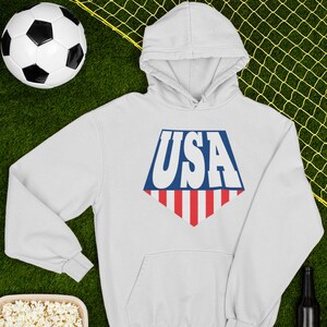 May include: White hooded sweatshirt with a blue and red USA graphic on the front. The graphic is in the shape of a shield with red and white stripes at the bottom.