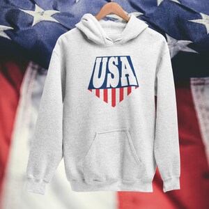 May include: A gray hooded sweatshirt with a white graphic of the word "USA" in a bold, blocky font. The graphic is in the shape of a shield with red and white stripes at the bottom, resembling the American flag.