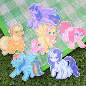 May include: A selection of cartoon pony stickers in various colours. The stickers depict different ponies in various poses, including pink, yellow, blue, purple, and orange. They are arranged on a green and white checkered picnic blanket.