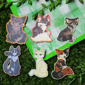 May include: Six die-cut stickers featuring illustrated cats in various colors and patterns. The collection includes a dark grey tabby, a black and white cat, a brown tabby, a blue-grey cat, a cream-colored cat, and a calico cat with orange and black.