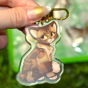 May include: A clear acrylic keychain featuring a stylized illustration of a brown tabby cat with large yellow eyes and a light green neck accessory. The cat is depicted in a sitting position with its tail wrapped around its body, attached to a gold-colored metal lobster clasp.