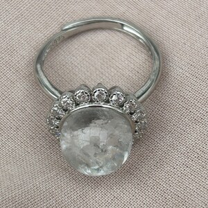 Peut inclure: Une bague en argent avec une grosse pierre précieuse ronde et transparente au centre, entourée de petites pierres transparentes. L'anneau est fin et poli, avec l'inscription "S925". Le sertissage autour de la pierre principale a un motif festonné.