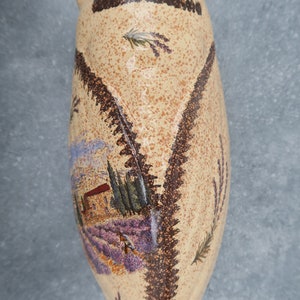 May include: A beige ceramic vase with a speckled finish and a painted scene of a lavender field and a house. The vase has a brown border and lavender sprig details.