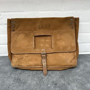 Peut inclure: Sac messager en toile marron avec un rabat rectangulaire et une fermeture à boucle en cuir. Le sac porte les lettres "SNCF" et comporte des rivets métalliques sur les côtés. Le sac est posé sur une surface grise.