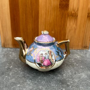 Peut inclure: Petite théière en porcelaine ornée, avec une anse, un bec et des bordures dorés. Le corps est bleu clair avec une scène décorative de personnages en costumes d'époque. Le couvercle est lavande et la base est blanche.