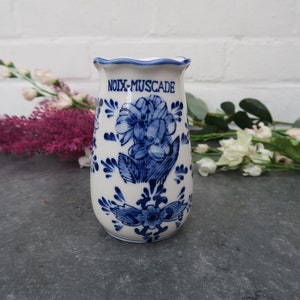 Może przedstawiać: Biały ceramiczny wazon z niebieskim kwiatowym wzorem i napisem "NOIX-MUSCADE". Wazon ma falisty brzeg i jest ozdobiony niebieskimi kwiatami i liśćmi. Wazon ma około 15 cm wysokości.