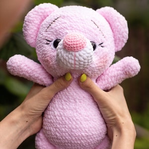 May include: A pink crocheted teddy bear with white accents on the nose and feet. The bear has black eyes and a pink nose. The bear is being held by a person's hands.