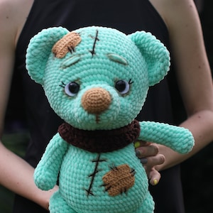 May include: A teal blue crocheted teddy bear with brown yarn accents. The bear has a stitched patch on its head and a brown yarn scarf around its neck.