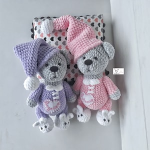 May include: Two crocheted teddy bears wearing pajamas and sleeping caps. The bear on the left is wearing a lavender sleeping cap and pajamas, while the bear on the right is wearing a pink sleeping cap and pajamas. Both bears have gray fur and are lying on a white surface.