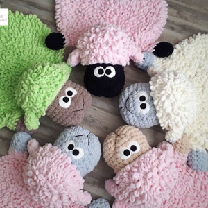 May include: Five crocheted sheep rugs in different colors, including green, pink, white, gray, and brown. Each rug has a fluffy texture and a cute sheep face with black eyes and a black nose.