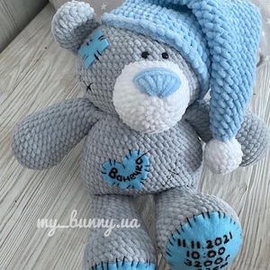 May include: A gray crocheted teddy bear wearing a blue crocheted hat. The bear has a white face with a blue nose and black eyes. The bear has a blue heart-shaped patch on its chest with the word "Ванечка" embroidered on it. The bear's feet are blue and have the date "11.11.2021" and the time "10:00" embroidered on them.