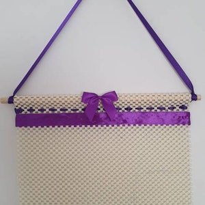 May include: A cream-coloured woven wall hanging with a grid pattern. It has a wooden dowel, purple ribbon accents, and a decorative bow. The hanging is suspended by a purple ribbon.