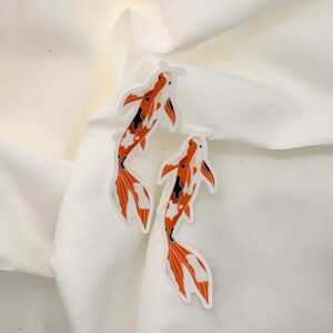May include: Two orange and white koi fish stickers on a white background. The fish are facing different directions and have black markings.