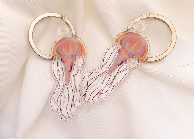 May include: Two clear acrylic keychains featuring a pink and orange jellyfish design. The jellyfish have long, flowing tentacles.