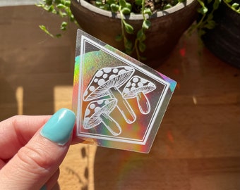 Może przedstawiać: Holograficzna naklejka przedstawiająca białe kontury trzech grzybów z białymi kropkami. Naklejka jest tęczowa i ma tęczowy połysk.