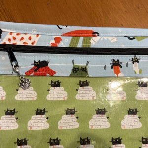 K&ouml;nnte beinhalten: Eine Stofftasche mit schwarzem Rei&szlig;verschluss und einem Pfotenanh&auml;nger. Die Tasche hat eine hellblaue Oberseite mit Katzenmuster und eine gr&uuml;ne Unterseite mit einem Katzen- und Deckenmuster. Die Tasche ist f&uuml;r die Aufbewahrung konzipiert.