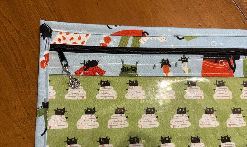 K&ouml;nnte beinhalten: Eine Stofftasche mit schwarzem Rei&szlig;verschluss und einem Pfotenanh&auml;nger. Die Tasche hat eine hellblaue Oberseite mit Katzenmuster und eine gr&uuml;ne Unterseite mit einem Katzen- und Deckenmuster. Die Tasche ist f&uuml;r die Aufbewahrung konzipiert.