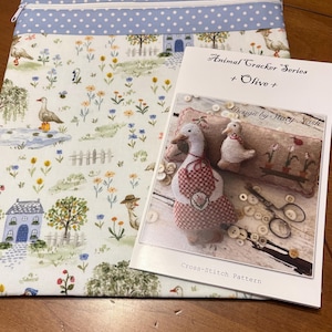 May include: A fabric case with a blue polka dot top and a zipper, next to a cross-stitch pattern. The pattern features a goose design with the text "Animal Cracker Series + Olive + Design by Stacy Nash" and the words "Cross-Stitch Pattern".