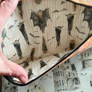K&ouml;nnte beinhalten: Eine Halloween-Rei&szlig;verschlusstasche mit schwarzem Rei&szlig;verschluss und orangefarbener Einfassung. Die Innenseite zeigt einen Aufdruck von Flederm&auml;usen und Vampirfiguren auf gestreiftem Hintergrund. Die Au&szlig;enseite zeigt ein Muster aus Spukh&auml;usern und Flederm&auml;usen.