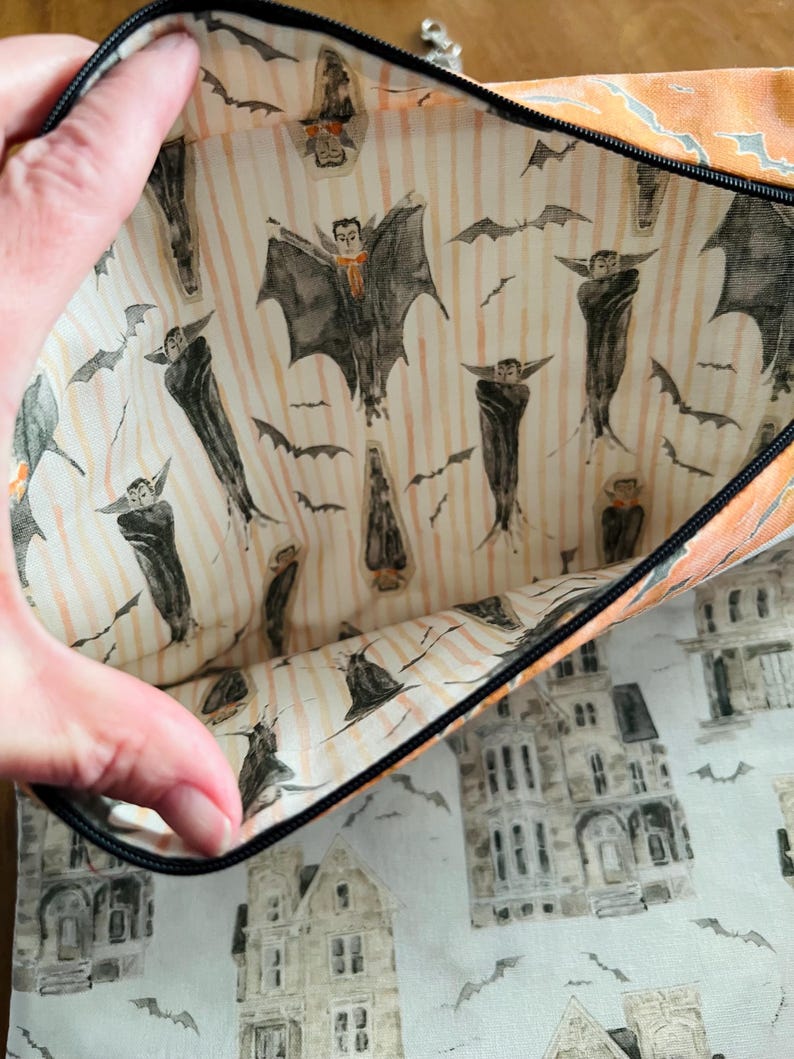 K&ouml;nnte beinhalten: Eine Halloween-Rei&szlig;verschlusstasche mit schwarzem Rei&szlig;verschluss und orangefarbener Einfassung. Die Innenseite zeigt einen Aufdruck von Flederm&auml;usen und Vampirfiguren auf gestreiftem Hintergrund. Die Au&szlig;enseite zeigt ein Muster aus Spukh&auml;usern und Flederm&auml;usen.