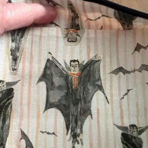 K&ouml;nnte beinhalten: Nahaufnahme eines Halloween-Stoffs mit einem sich wiederholenden Muster aus Aquarell-Vampiren und Flederm&auml;usen. Die Vampire haben dunkle Umh&auml;nge und orangefarbene Fliegen. Der Hintergrund hat vertikale Pfirsichstreifen.