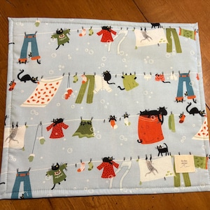 K&ouml;nnte beinhalten: Hellblaues Platzset mit einem skurrilen Design aus W&auml;scheleinen, h&auml;ngender Kleidung und schwarzen Katzen. Die Kleidung umfasst Hosen, Kleider, Hemden und Socken. Das Platzset hat einen gen&auml;hten Rand und ein kleines Etikett in der unteren rechten Ecke.