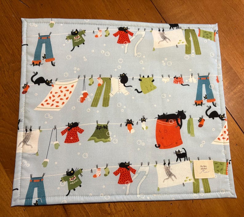 K&ouml;nnte beinhalten: Hellblaues Platzset mit einem skurrilen Design aus W&auml;scheleinen, h&auml;ngender Kleidung und schwarzen Katzen. Die Kleidung umfasst Hosen, Kleider, Hemden und Socken. Das Platzset hat einen gen&auml;hten Rand und ein kleines Etikett in der unteren rechten Ecke.