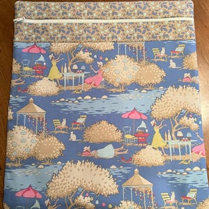 May include: A blue fabric zippered pouch with a whimsical garden scene print. The print features figures in dresses, cats, tables, and umbrellas. The top border is a floral pattern in tan and blue. The zipper is silver.