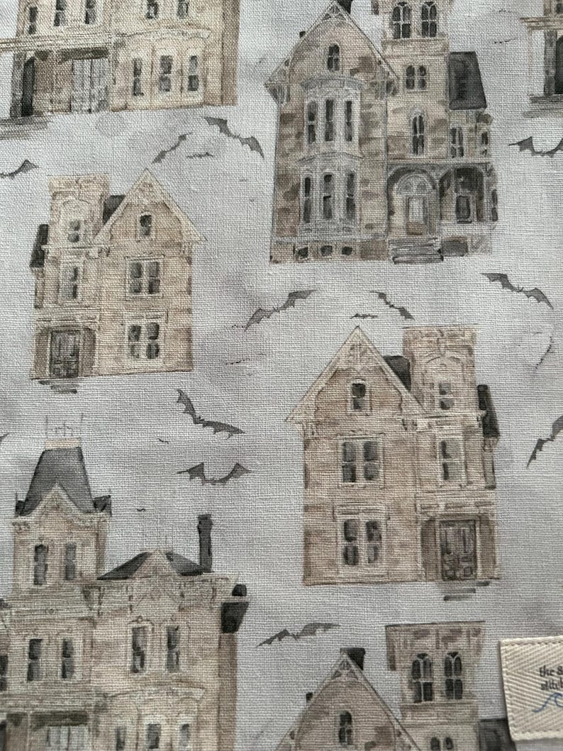 K&ouml;nnte beinhalten: Stoff mit einem sich wiederholenden Muster von Vintage-H&auml;usern mit Spuk-Optik in Beige-, Grau- und Braunt&ouml;nen. Silhouetten von Flederm&auml;usen sind &uuml;ber das Design verstreut. Die H&auml;user haben mehrere Fenster, T&uuml;ren und architektonische Details.