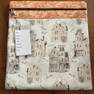 K&ouml;nnte beinhalten: Eine rechteckige Stofftasche mit einem schwarzen Rei&szlig;verschluss und einem silberfarbenen Rei&szlig;verschlussanh&auml;nger. Die Tasche hat einen cremefarbenen Hintergrund mit Illustrationen von Spukh&auml;usern und Flederm&auml;usen. Der obere Teil ist orange mit einem Fledermausmuster.