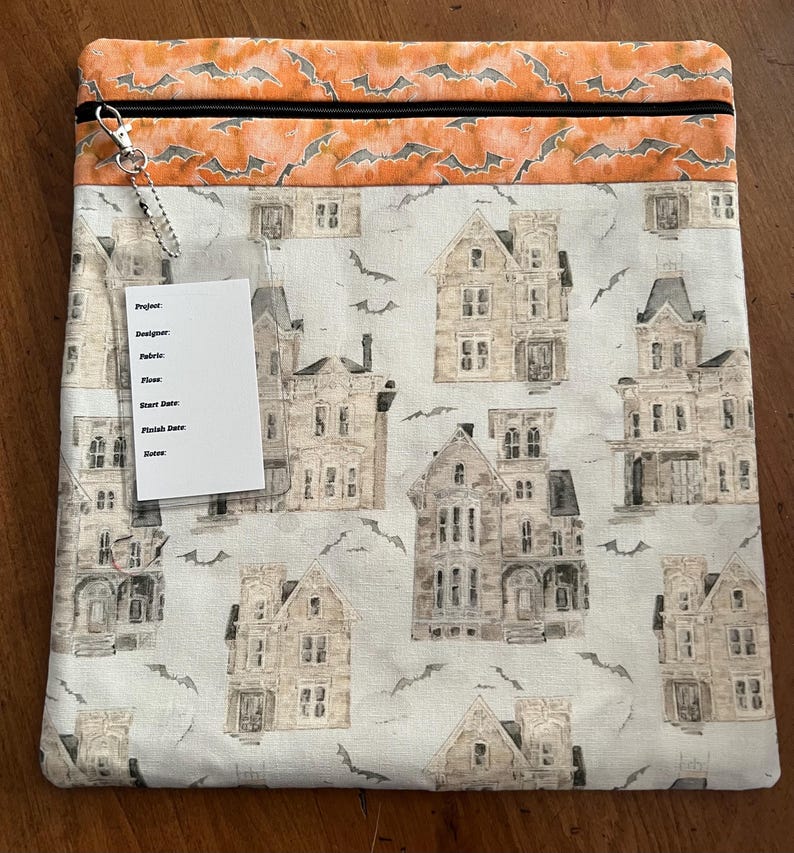 K&ouml;nnte beinhalten: Eine rechteckige Stofftasche mit einem schwarzen Rei&szlig;verschluss und einem silberfarbenen Rei&szlig;verschlussanh&auml;nger. Die Tasche hat einen cremefarbenen Hintergrund mit Illustrationen von Spukh&auml;usern und Flederm&auml;usen. Der obere Teil ist orange mit einem Fledermausmuster.