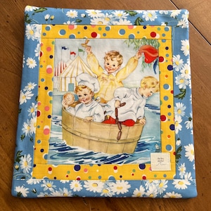 May include: A quilted fabric item with a vintage illustration of three children in a boat. The border is blue with white daisies, a yellow polka dot stripe, and a yellow border. The item has a small white tag with text.