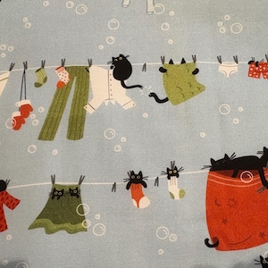K&ouml;nnte beinhalten: Hellblauer Stoff mit einem skurrilen Muster von Kleidung, die an einer W&auml;scheleine h&auml;ngt, mit schwarzen Katzen. Die Kleidung umfasst Kleider, Hosen, Socken und eine Decke. Der Hintergrund hat wei&szlig;e Blasen.