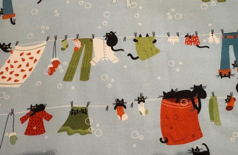 K&ouml;nnte beinhalten: Hellblauer Stoff mit einem skurrilen Muster von Kleidung, die an einer W&auml;scheleine h&auml;ngt, mit schwarzen Katzen. Die Kleidung umfasst Kleider, Hosen, Socken und eine Decke. Der Hintergrund hat wei&szlig;e Blasen.