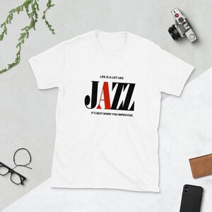 Puede incluir: Una camiseta blanca con el texto "LIFE IS A LOT LIKE JAZZ. IT'S BEST WHEN YOU IMPROVISE." en negro y rojo. La camiseta se muestra sobre una superficie clara con una cámara, gafas y una cartera.