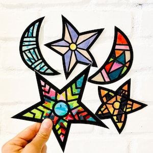 May include: Five colorful paper cutouts of stars and crescent moons. The stars are in shades of blue, purple, and yellow. The moons are in shades of blue, green, and red. The cutouts are on a white brick background.