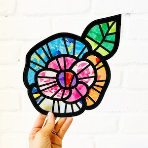 May include: A colourful paper flower with a black outline and a green leaf. The flower is made of different coloured paper squares that are layered on top of each other. The flower is a vibrant and cheerful piece of art.