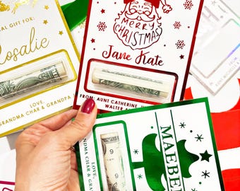 May include: Christmas gift cards with rolled-up dollar bills. One card has a gold foil design with the name "Rosalie". Another card has a Santa Claus design with the text "Merry Christmas Jane Kate". A third card has a green Christmas tree design with the name "Maebel".