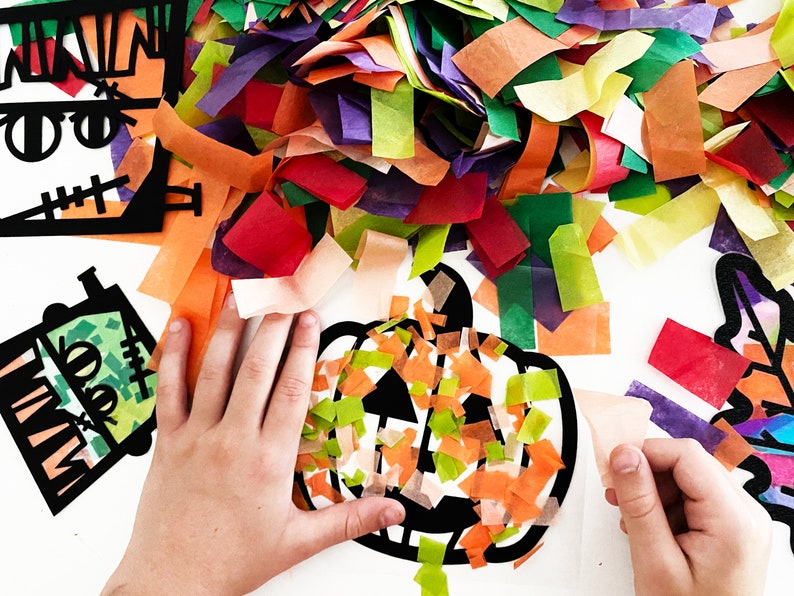 May include: A child's hands are placing colorful tissue paper onto a black paper pumpkin cutout. The tissue paper is in shades of orange, green, and yellow.