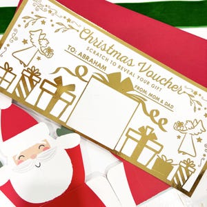 May include: A Christmas voucher with gold foil accents, featuring the text "Christmas Voucher" and "Scratch to Reveal Your Gift". The voucher is addressed "TO: ABRAHAM" and signed "FROM: MOM & DAD". A Santa Claus card is visible in the foreground.