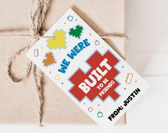May include: A white gift tag with a red, blue, and yellow Lego-themed design. The text on the tag reads "We were built to be friends" and "From: Justin".
