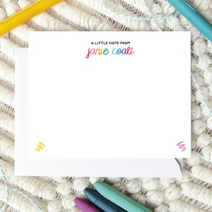 May include: White stationery set with a matching envelope. The stationery features the text "A LITTLE NOTE FROM janie coalt" in rainbow colors. Colorful scribbles are in the corners. Stationery is on a white textured surface.