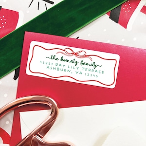 May include: A red envelope with a white label featuring the text "the komatz family" in a script font, with address details below. A green ribbon and patterned paper are in the background. A rose gold paper clip is in the foreground.