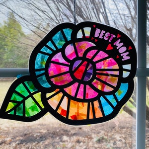 May include: A colorful paper craft flower with a leaf, decorated with tissue paper and the words "BEST MOM" in pink letters with small hearts.