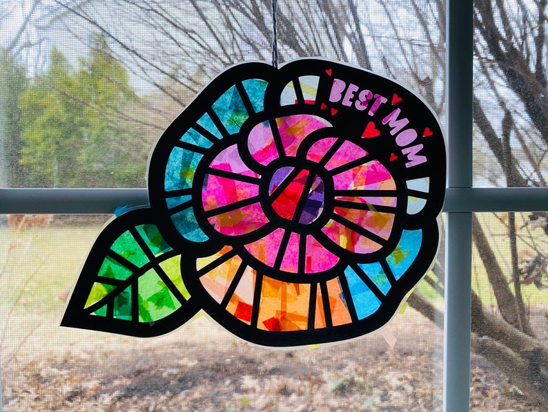 May include: A colorful paper craft flower with a leaf, decorated with tissue paper and the words "BEST MOM" in pink letters with small hearts.