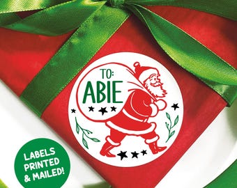 May include: A red wrapped present with a green ribbon and a white circular sticker. The sticker features a red Santa Claus carrying a sack, the text "TO: ABIE" in green, and black stars. A green circle with the text "LABELS PRINTED & MAILED!" is in the lower left.