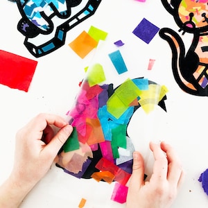 May include: A black cat silhouette is being decorated with colorful tissue paper. The tissue paper is being placed on top of the cat silhouette, which is on a white surface.