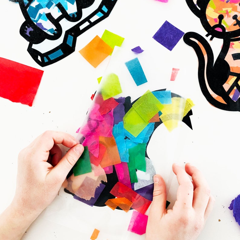 May include: A black cat silhouette is being decorated with colorful tissue paper. The tissue paper is being placed on top of the cat silhouette, which is on a white surface.
